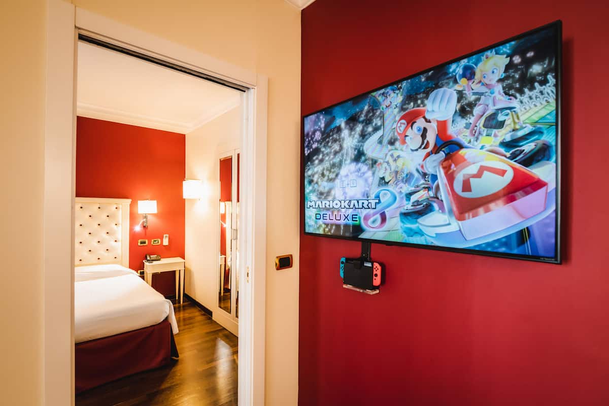 A hotel room with a red wall, a bed with a white headboard, a small bedside table with a lamp, and a television displaying a Mario Kart game.