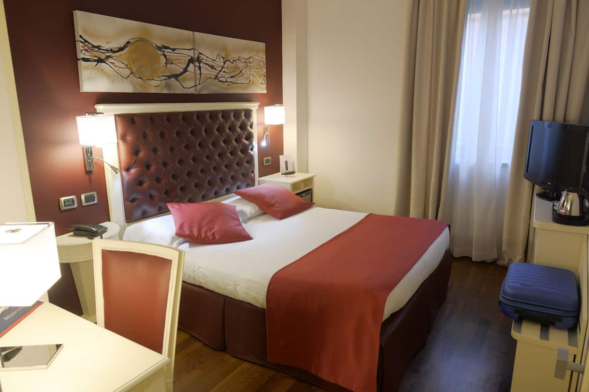 A hotel room with a large bed, a headboard, a desk with a lamp, a television, and a suitcase.