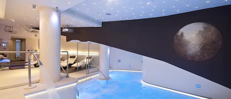 A modern indoor pool area with a large, spherical sculpture on the wall, a seating area with a glass balustrade, and a ceiling with recessed lighting and a projector.