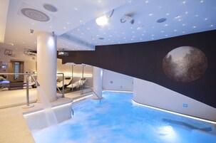 Un'area piscina moderna al chiuso, con una grande scultura sferica sulla parete, una zona relax con ringhiera in vetro, e un soffitto con illuminazione a incasso e un proiettore.