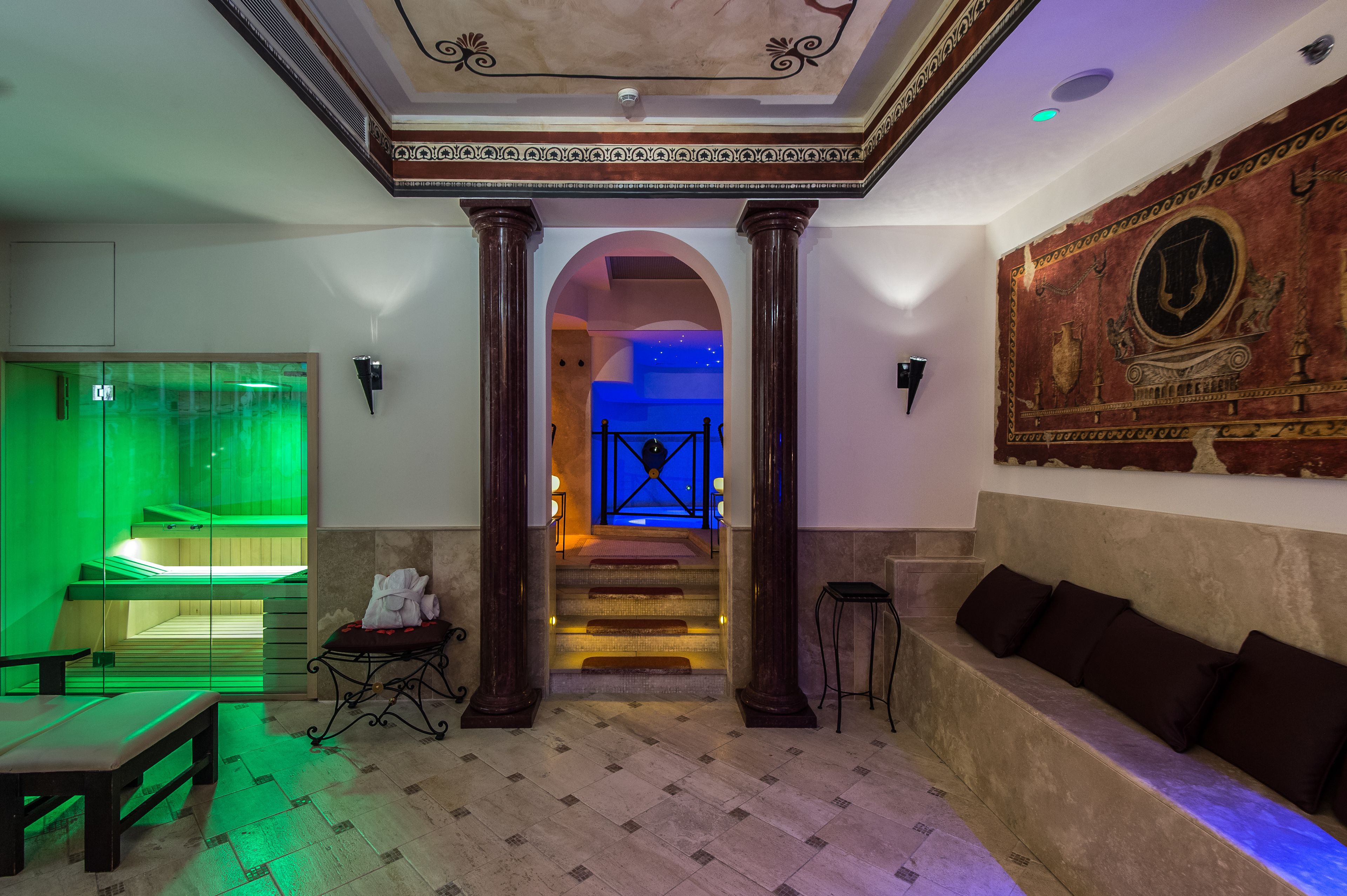 A lobby with a green-lit massage room, a staircase leading to a blue-lit area, and a seating area with a mural.