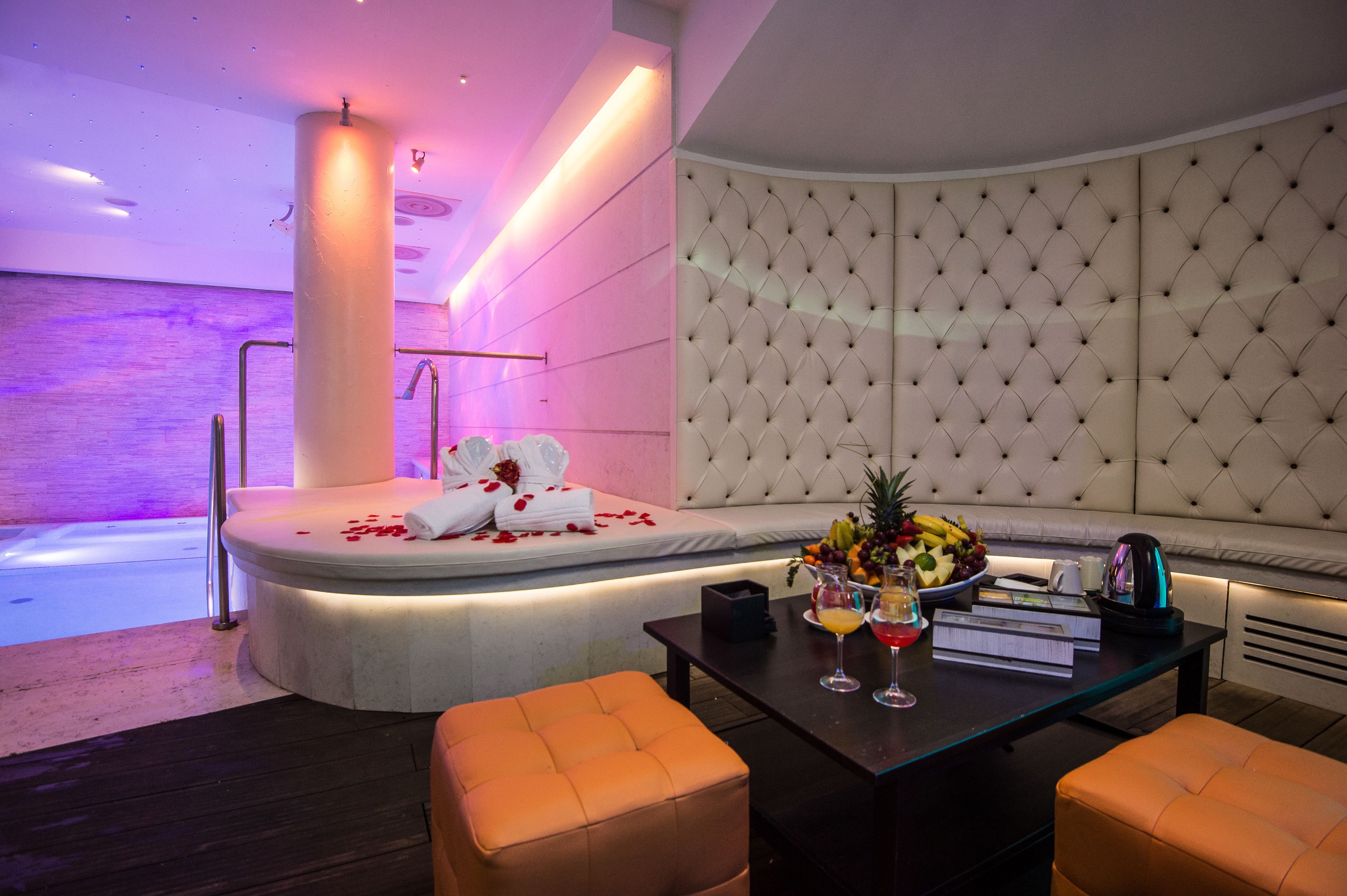 A modern hotel room with a plush sofa, a round table with a fruit bowl and drinks, and a large cushioned headboard.