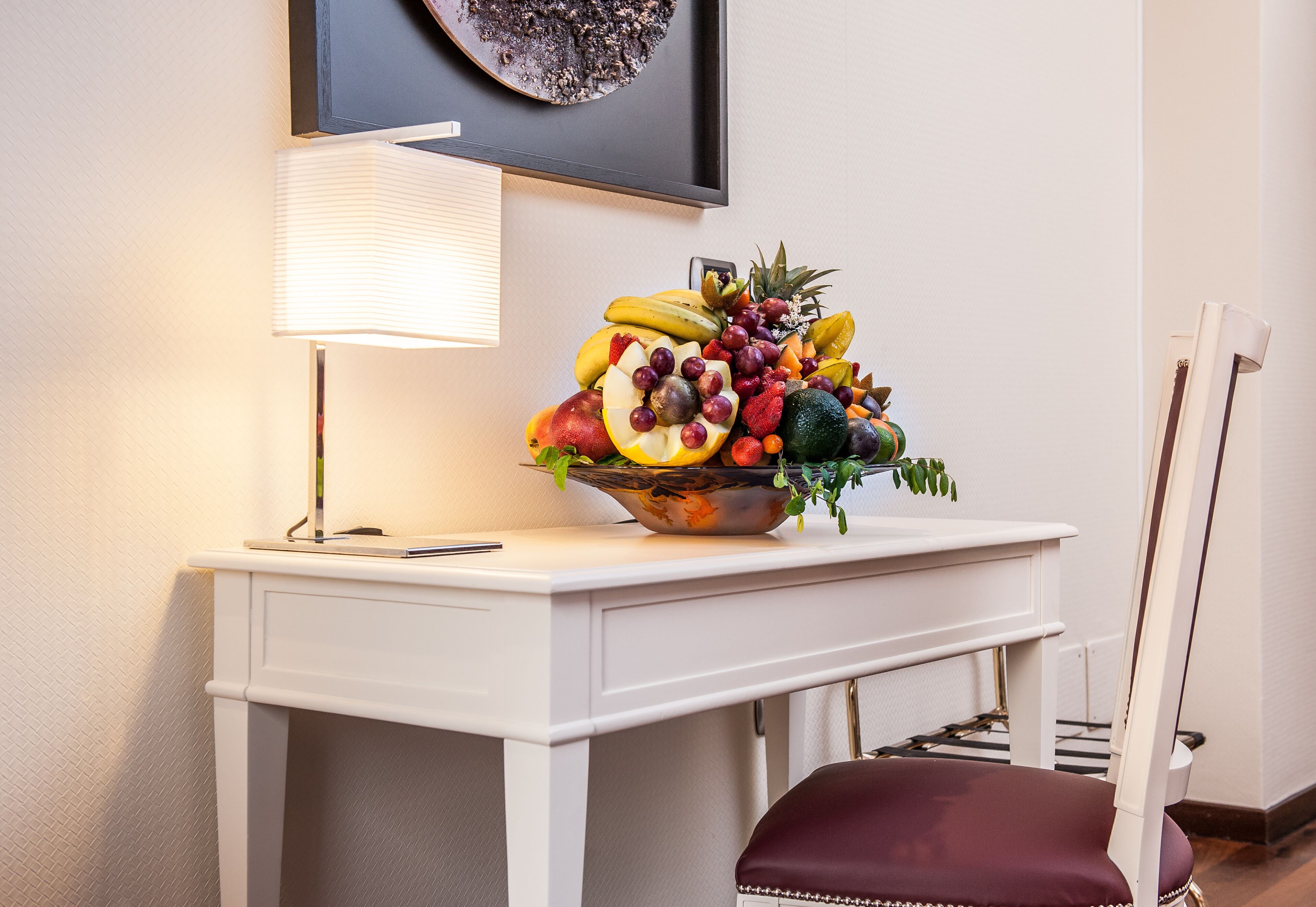 Et skrivebord med en skål frugt, en lampe og en stol.