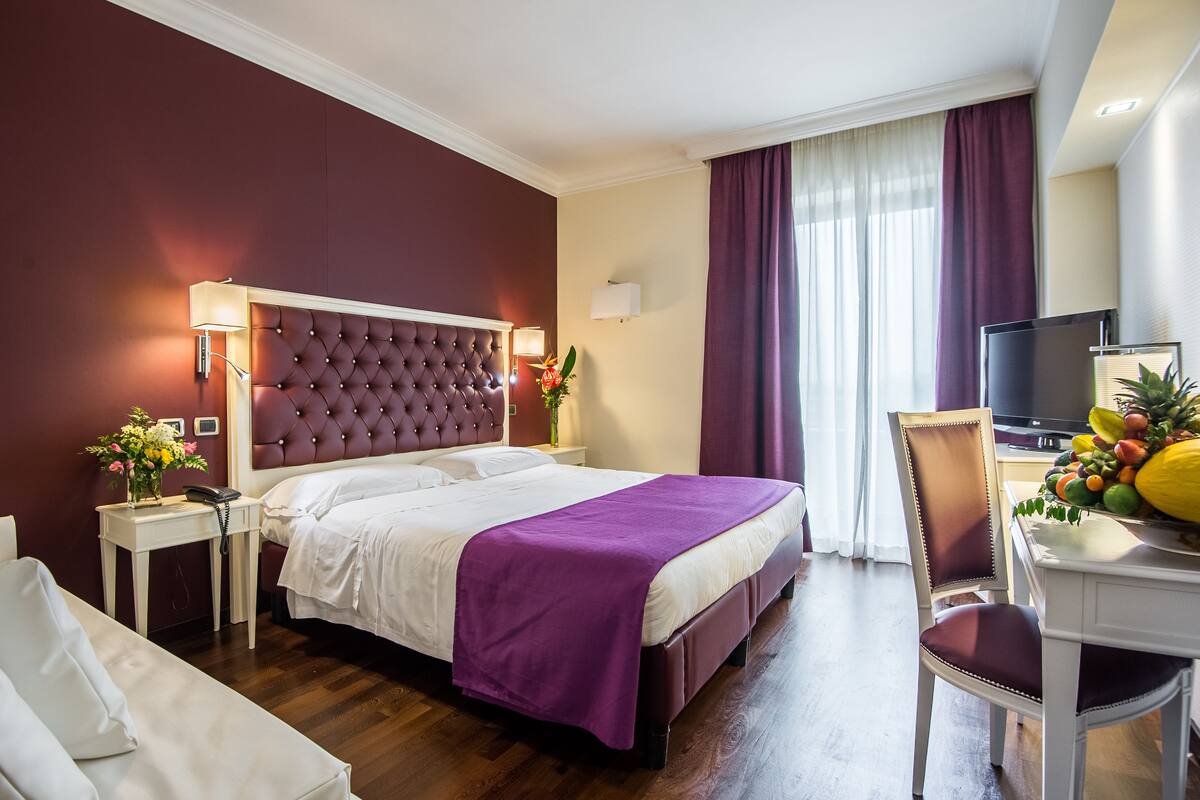 A hotel room with a large bed, a bedside table with a lamp, a TV, and a fruit bowl.