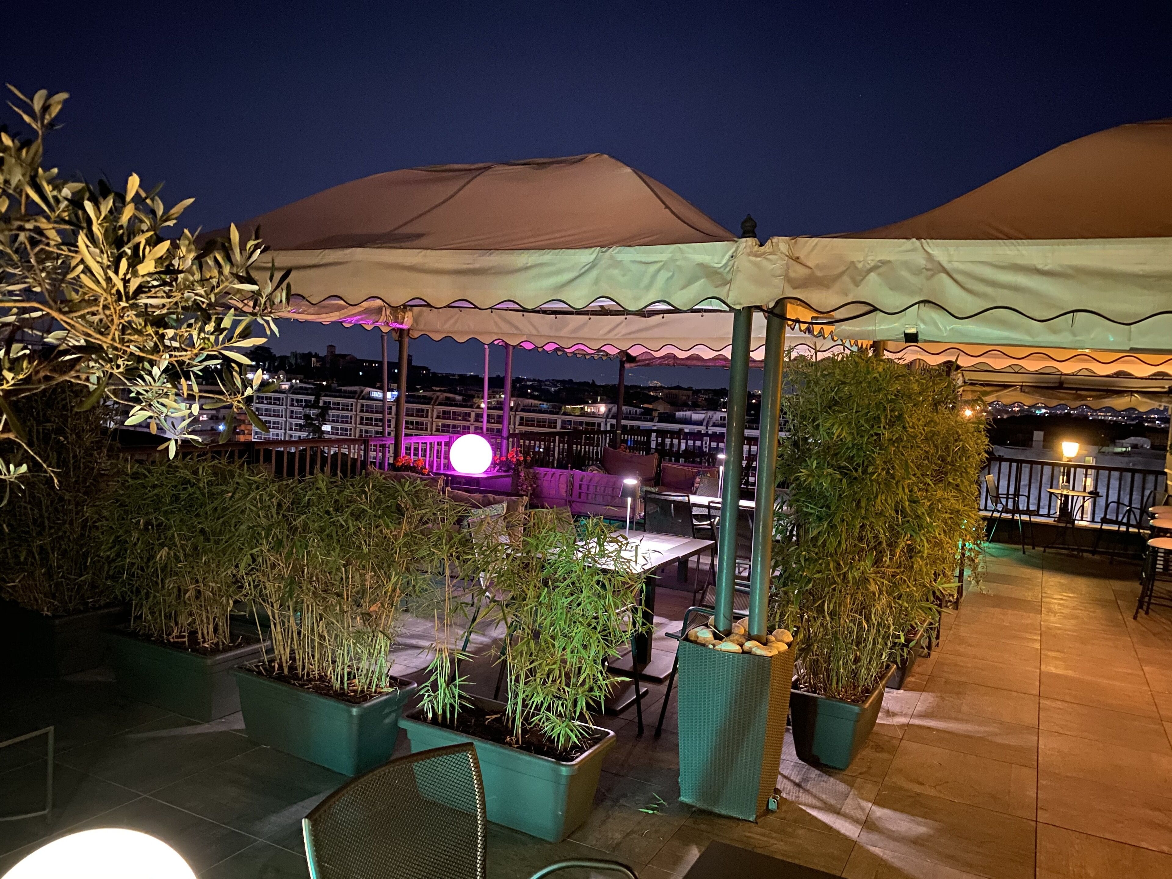 An outdoor patio area with tables and chairs, surrounded by potted plants and a large umbrella.