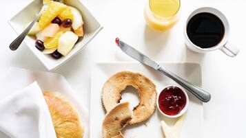 Une table dressée pour le petit-déjeuner avec un bol de fruits, un verre de jus d’orange, une tasse de café, un bagel tartiné de fromage frais et un croissant.