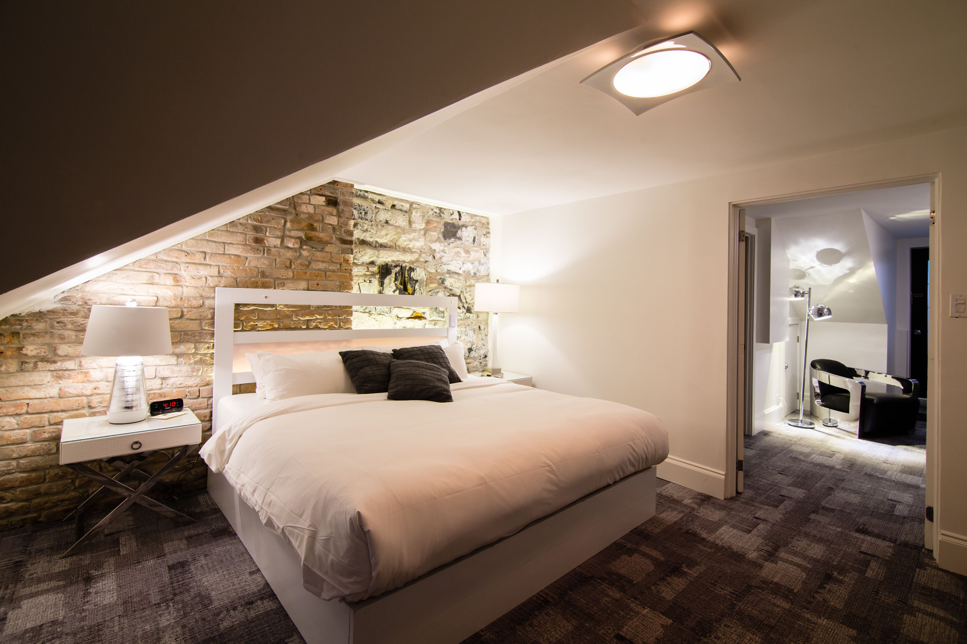 A bedroom with a stone wall, a bed with white bedding, a nightstand with a lamp, and a door leading to a bathroom.