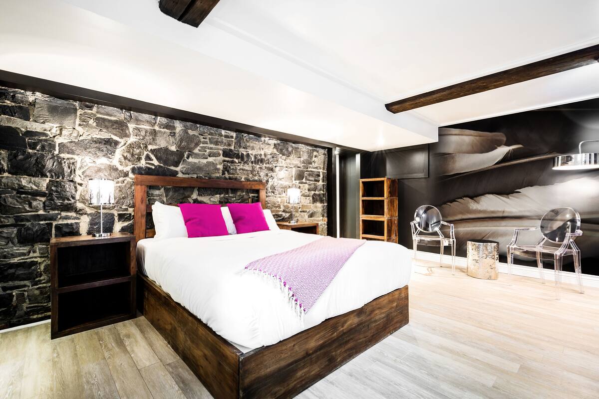 Une chambre avec un mur en pierre, un cadre de lit en bois, une couvre-lit blanche et un coussin rose.