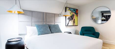 A modern bedroom with a large bed, two pendant lights, a round mirror, a teal armchair, and a colorful artwork on the wall.