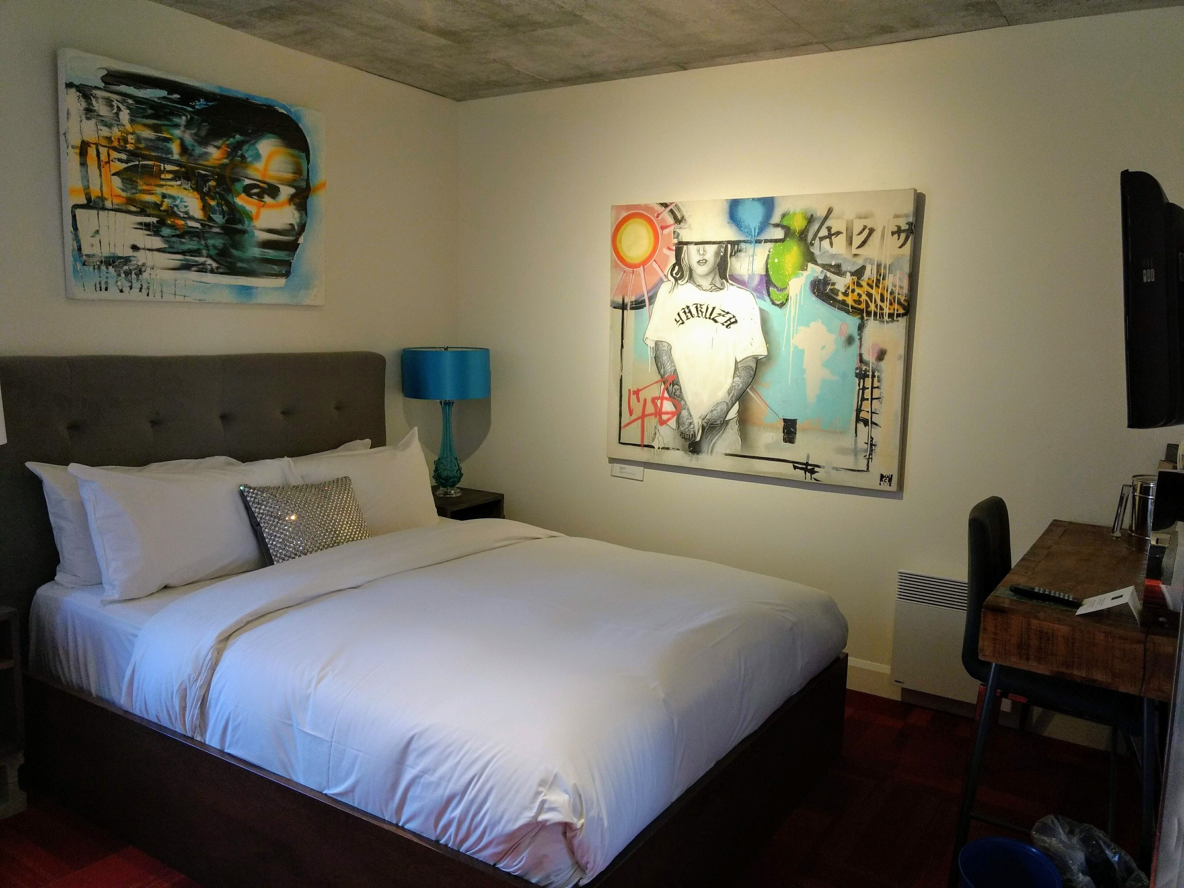 Una habitación de hotel con una cama grande, una mesita de noche con una lámpara, un escritorio con una silla y dos obras de arte en la pared.
