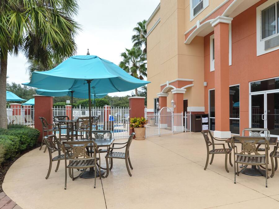 An outdoor patio with tables and chairs, a large blue umbrella, and a pool area in the background.