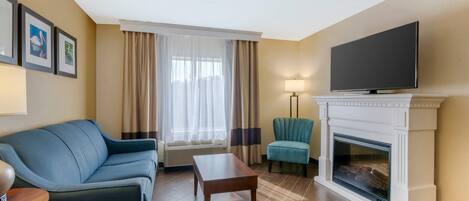 A hotel room with a sofa, a small table, a television, a fireplace, and a window with curtains.