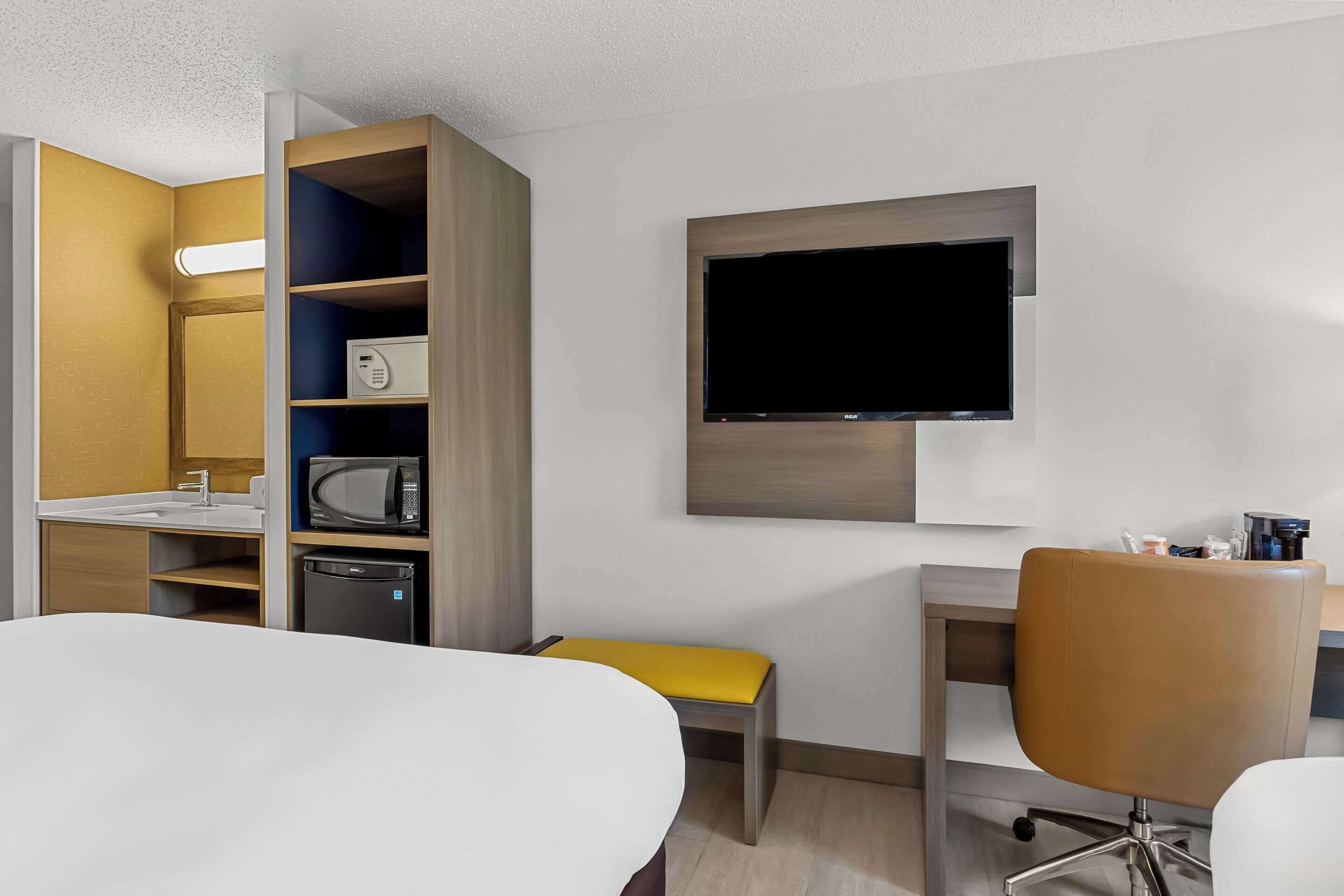 A hotel room with a bed, a desk, a chair, a TV mounted on the wall, a microwave, and a small cabinet.