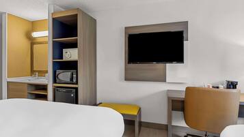 A hotel room with a bed, a desk, a chair, a TV mounted on the wall, a microwave, and a small cabinet.