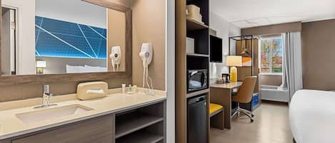 A hotel room with a large mirror, a sink, a microwave, a desk with a chair, and a bed.