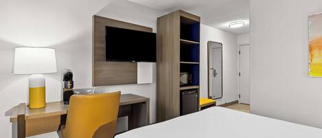 A hotel room with a bed, a desk with a lamp, a chair, a TV mounted on the wall, a microwave, and a small cabinet.