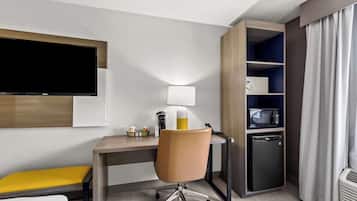 Habitación de hotel con escritorio, silla, televisor montado en la pared, lámpara, refrigerador miniatura, microondas y un pequeño calefactor.