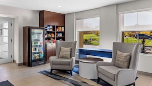 A modern hotel lobby with a refrigerator stocked with beverages, two grey armchairs, a round coffee table, and a large window offering a view of a green landscape.
