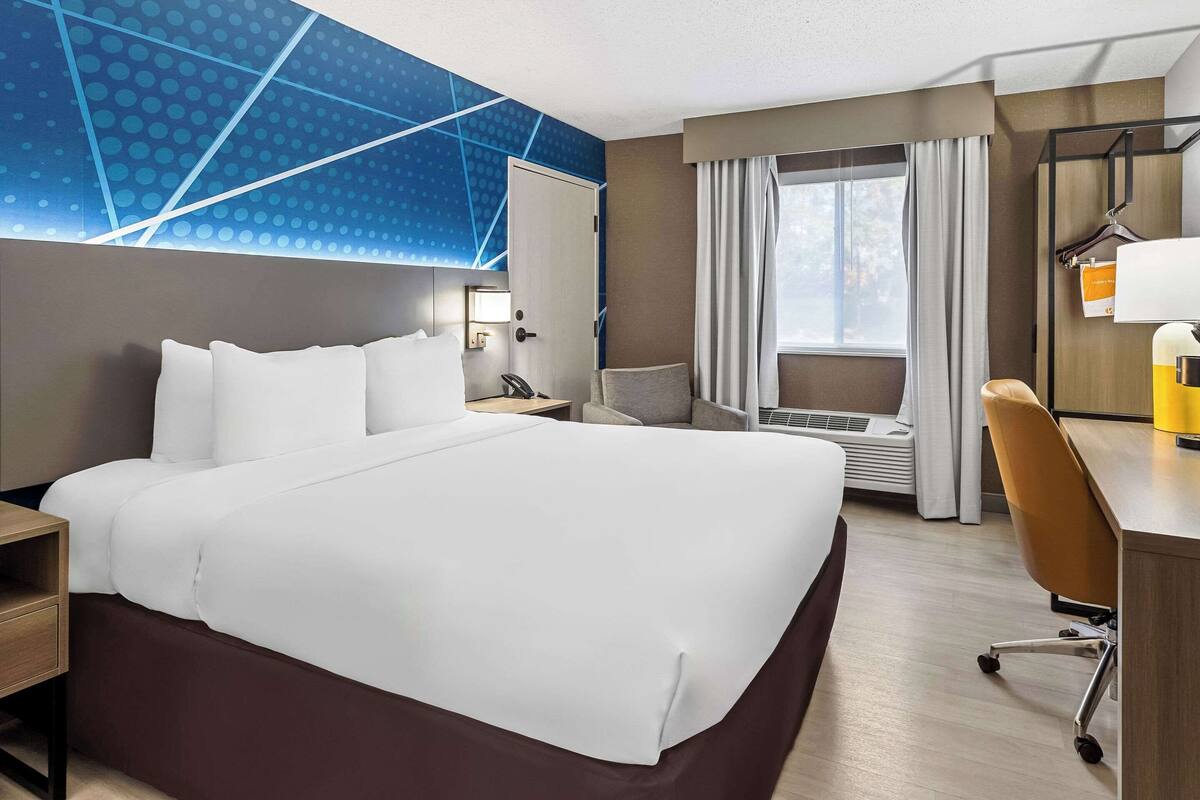 Habitación de hotel con una cama grande, un escritorio, una silla, una lámpara de escritorio, una ventana con cortinas y una pared con un diseño geométrico.