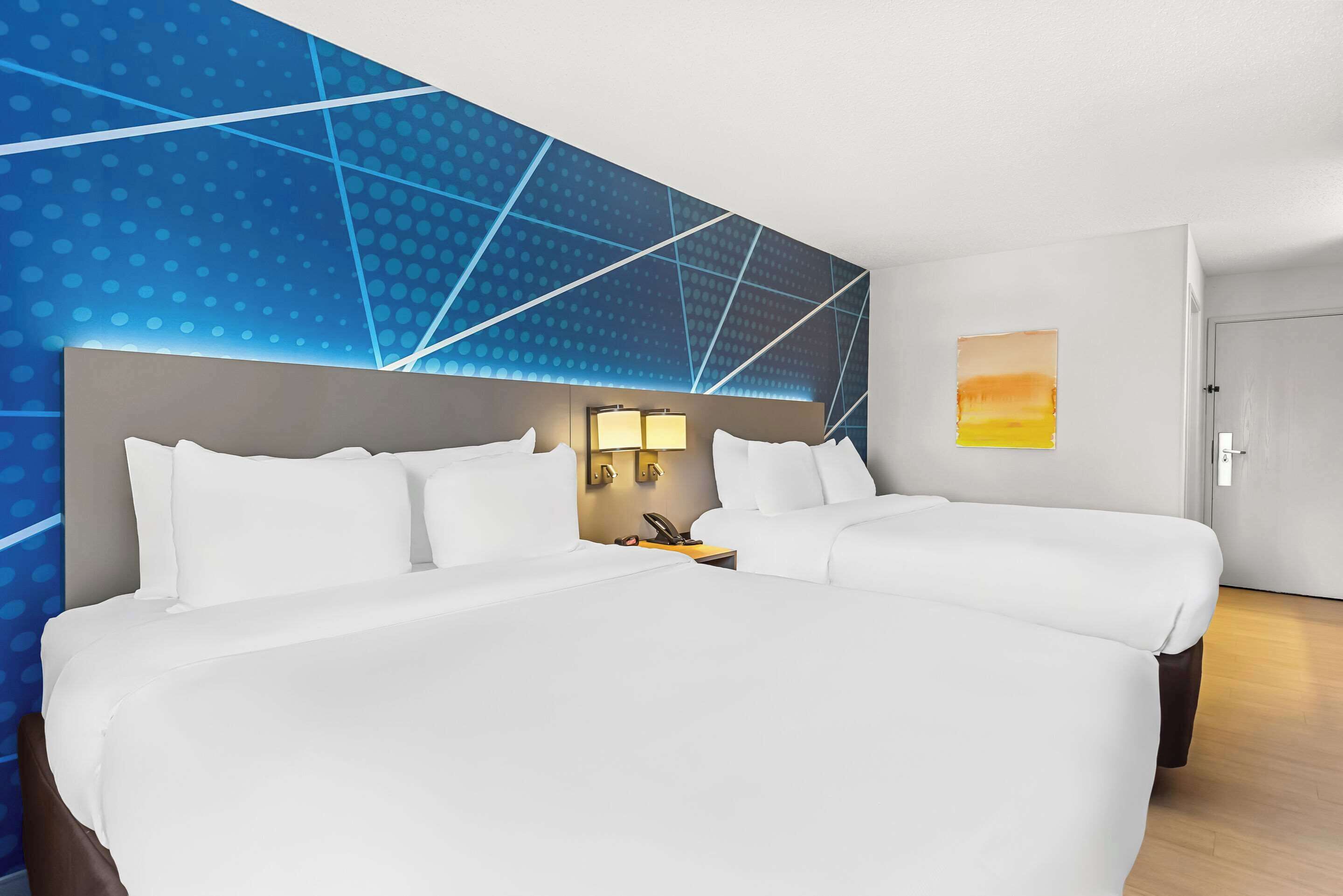 A hotel room with two beds, a large wall mural, a nightstand with a lamp, and a door.