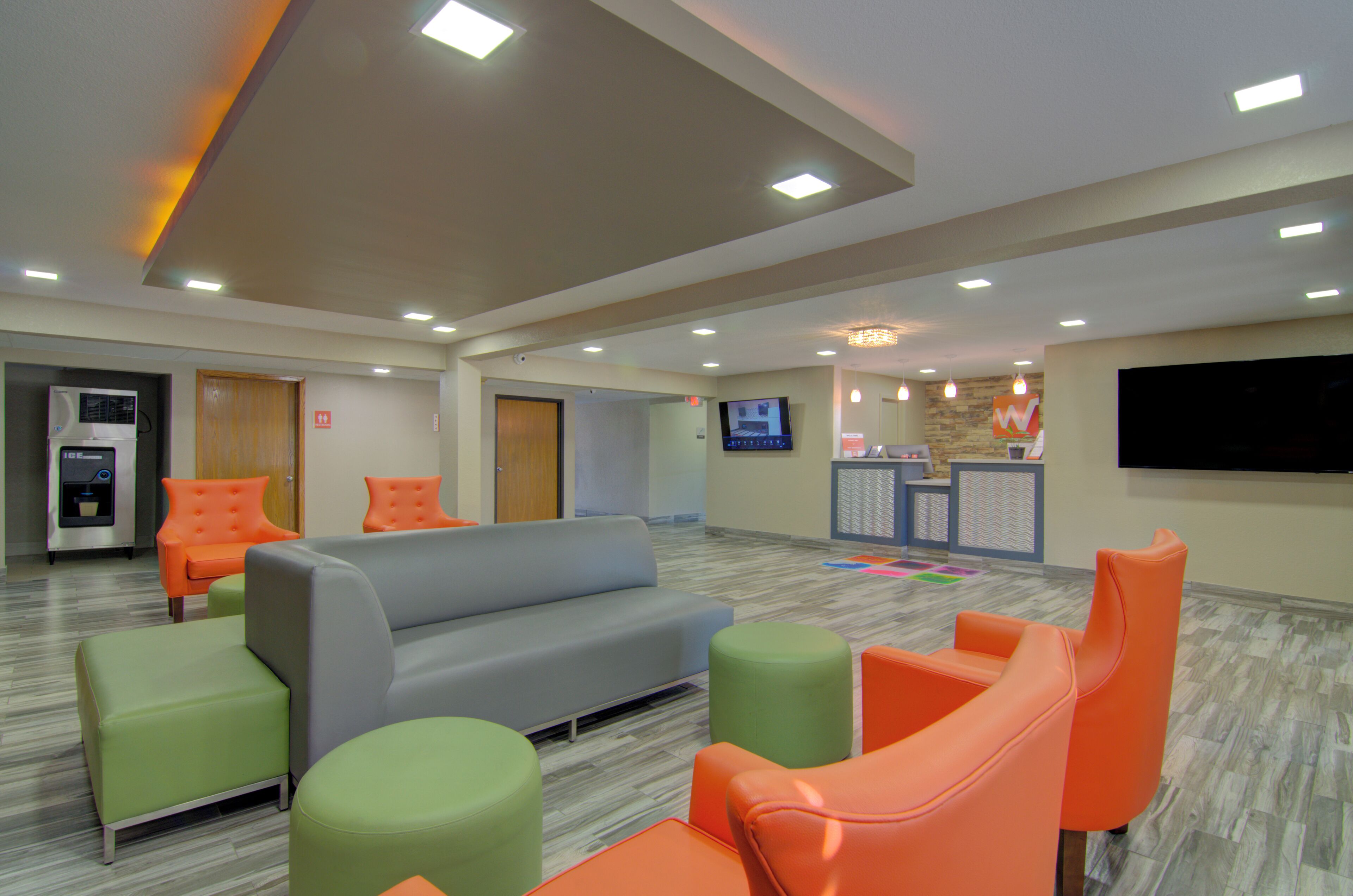 A spacious lobby with a grey sofa, orange and green chairs, a reception desk, and a TV mounted on the wall.