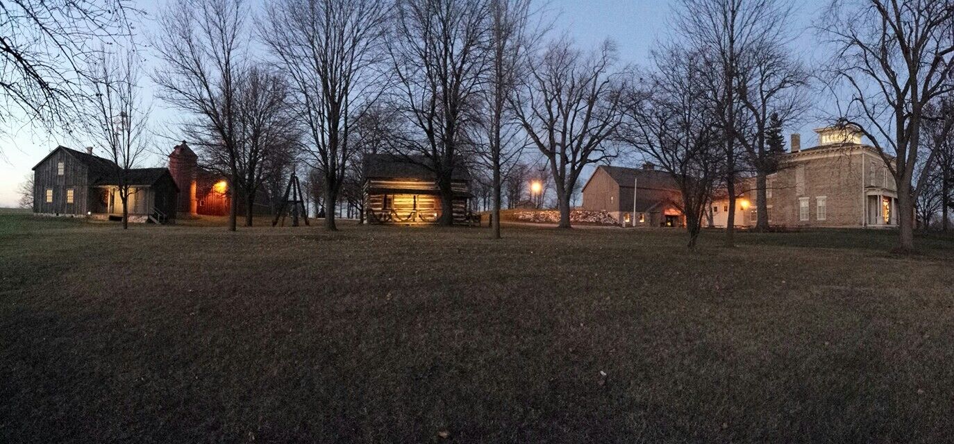 A historic building complex with multiple structures, including a large barn and a smaller log cabin, surrounded by open grassy fields and leafless trees.