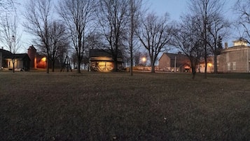 A historic building complex with multiple structures, including a large barn and a smaller log cabin, surrounded by open grassy fields and leafless trees.