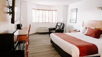 A hotel room with a bed, a desk with a chair, a TV, an air conditioner, and a window with blinds.