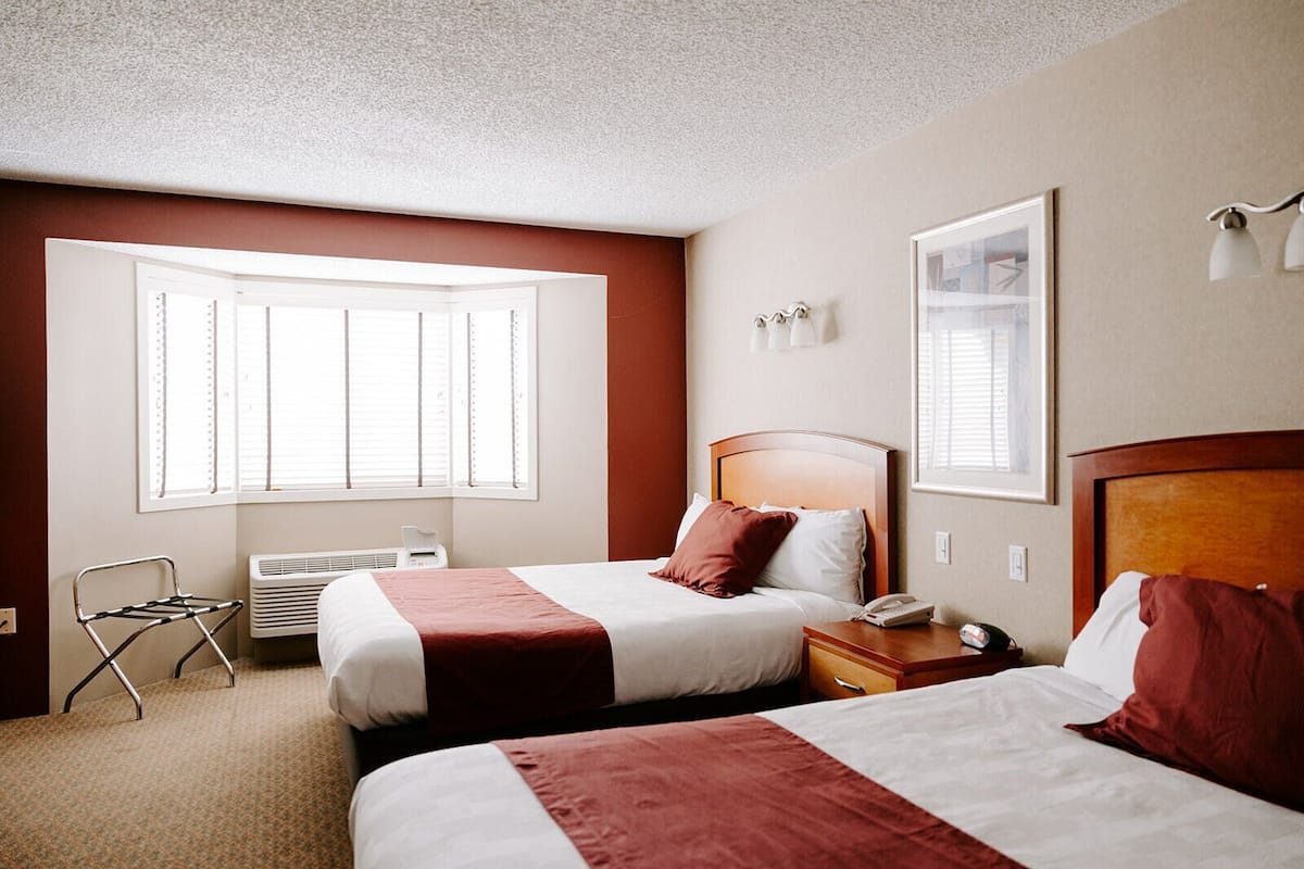 A hotel room with two beds, a window with blinds, a radiator, a bedside table with a lamp, and a framed picture on the wall.