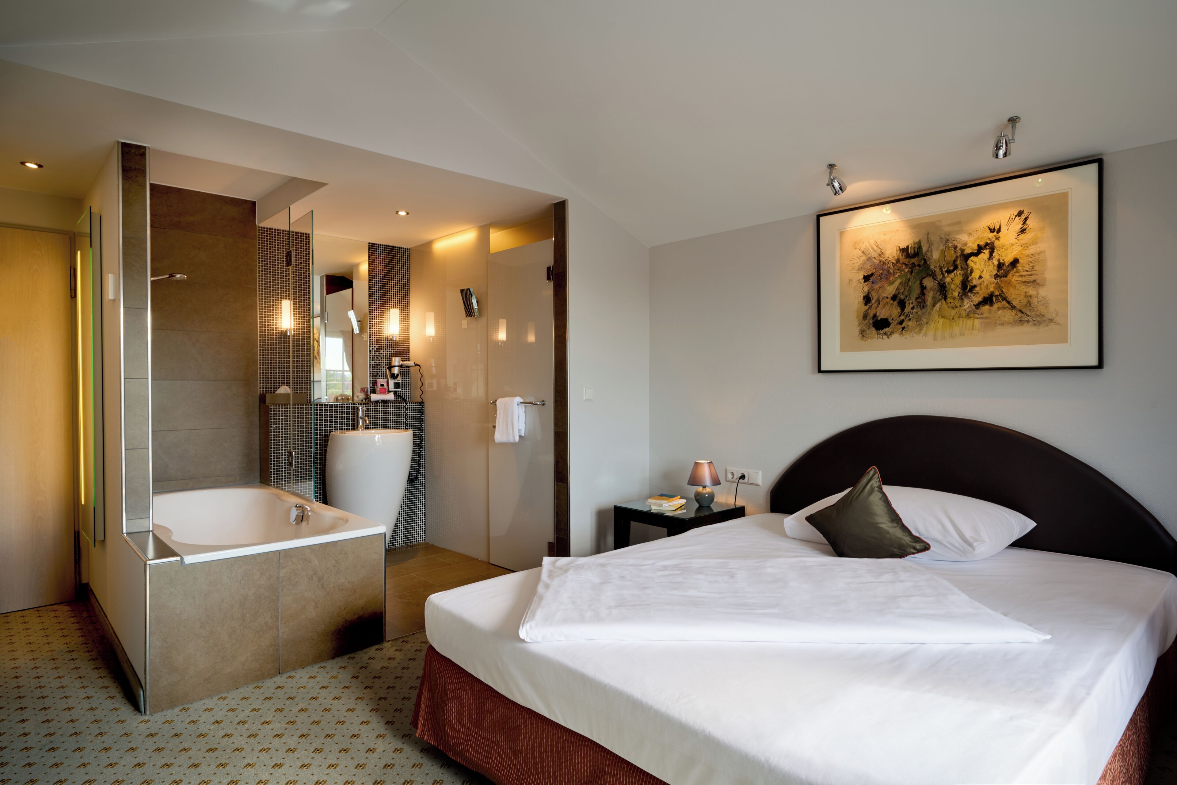 A hotel room with a large bed, a bathroom with a bathtub, and a painting on the wall.
