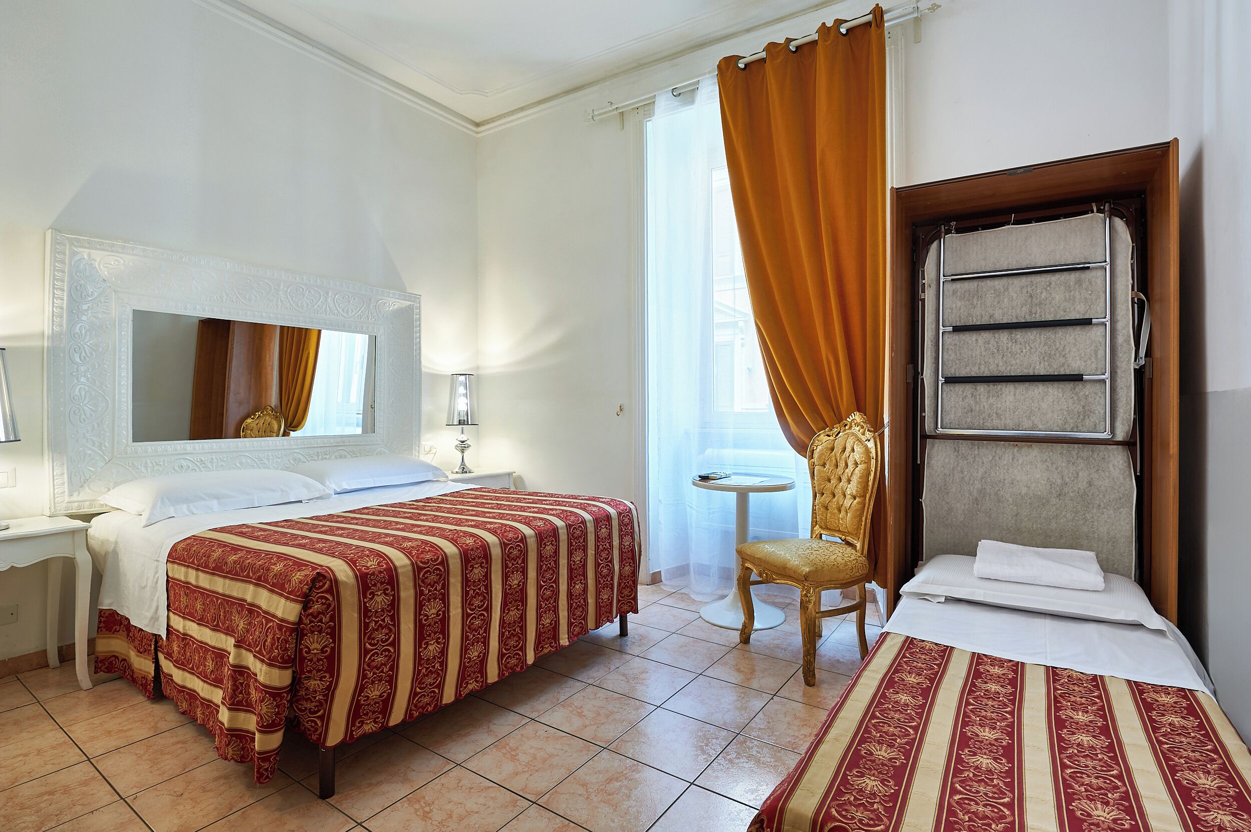 Camera d'albergo con due letti, una sedia, un tavolino, uno specchio e una finestra con tende.