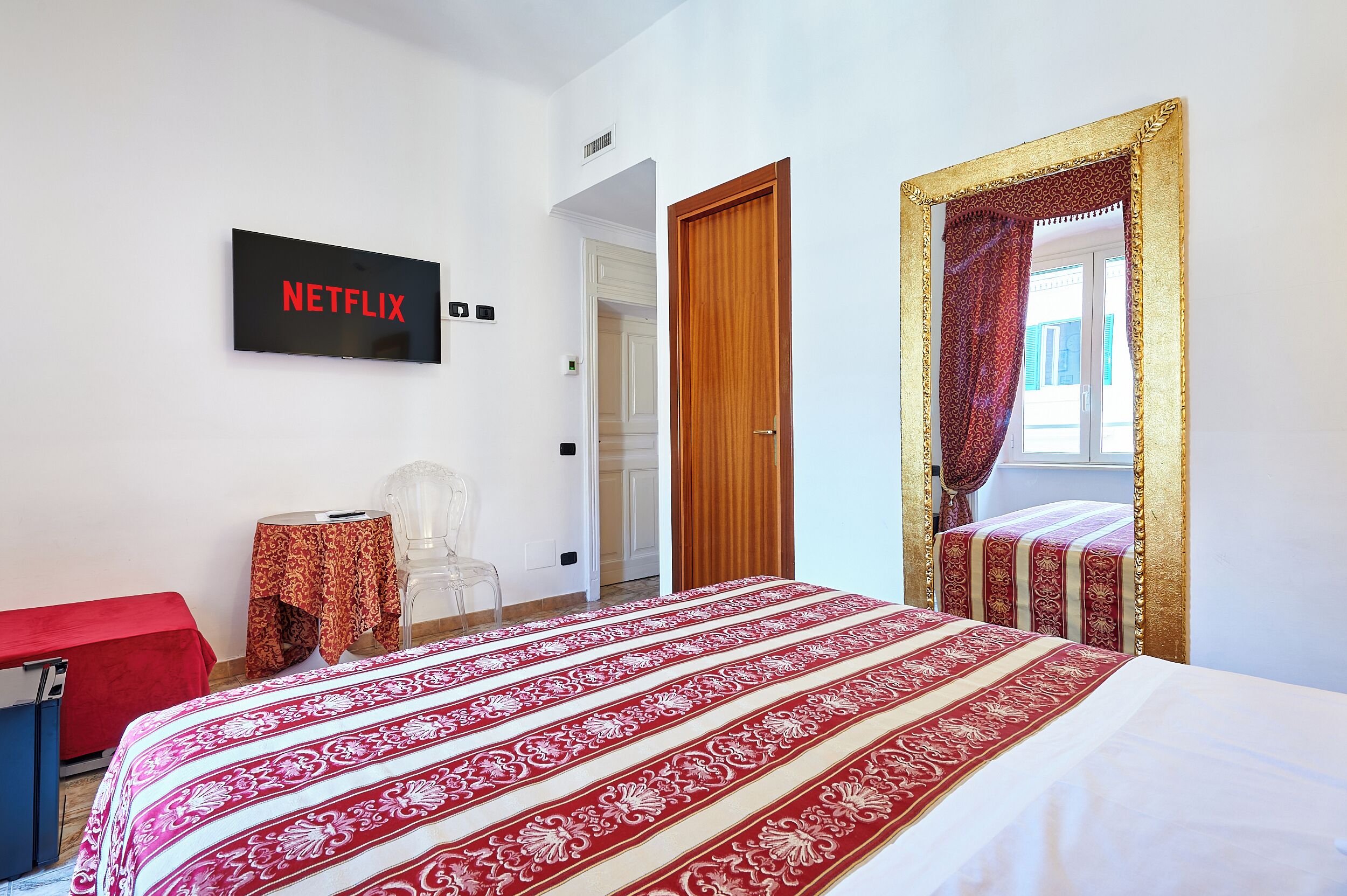 A hotel room with a bed, a TV mounted on the wall, a small table with a chair, and a door leading to another room.