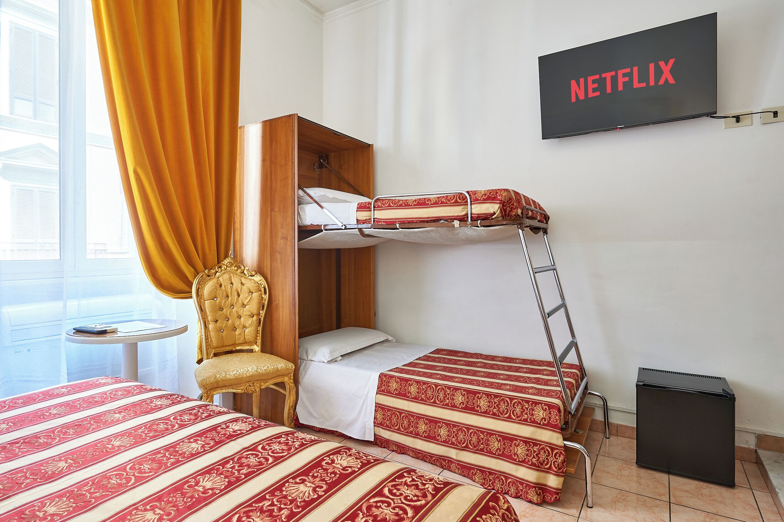A hotel room with a bunk bed, a TV mounted on the wall, a chair, a small table, and a window with curtains.