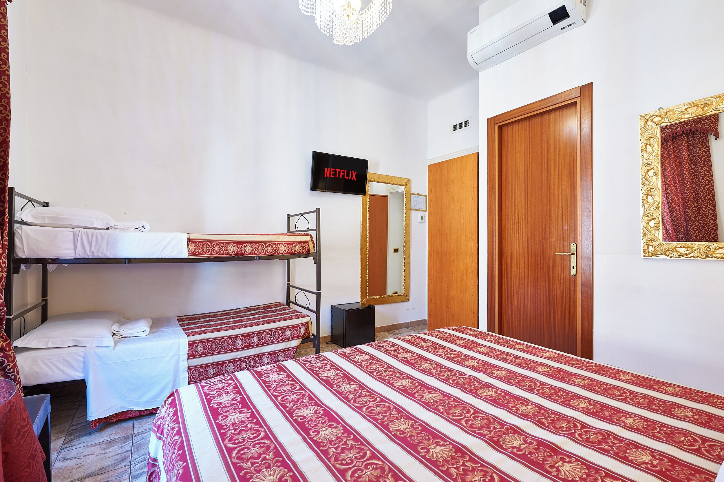 Habitación de hotel con dos camas literas, un televisor mostrando 'NETFLIX', una puerta de madera y un espejo con marco decorativo.