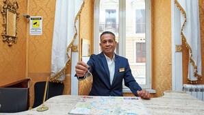 A man in a blue suit holding a key, standing in a room with a map on the table, a sign indicating a security area, and a view of buildings through a window. - Virginia Hotel (Rome)