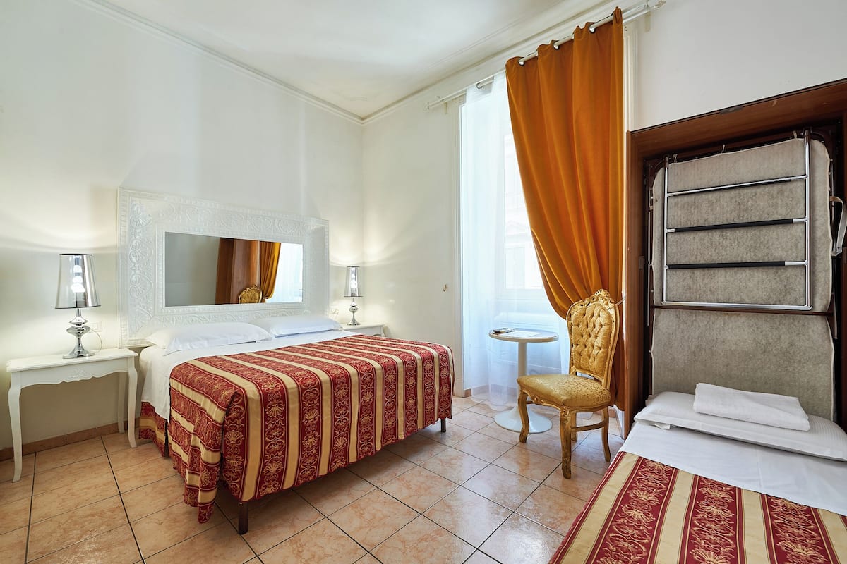 Une chambre d’hôtel avec un lit, une chaise, une petite table, un miroir et une fenêtre avec des rideaux.