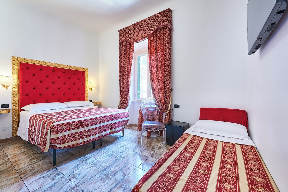 Une chambre d’hôtel avec deux lits, une tête de lit rouge et une fenêtre avec des rideaux rouges.