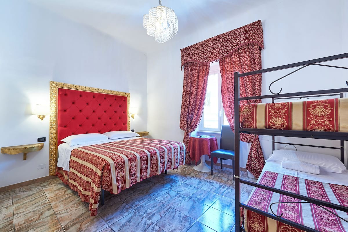 Une chambre d’hôtel avec un lit recouvert d’un tissu rouge, un lit superposé, un lustre et des rideaux à motifs.