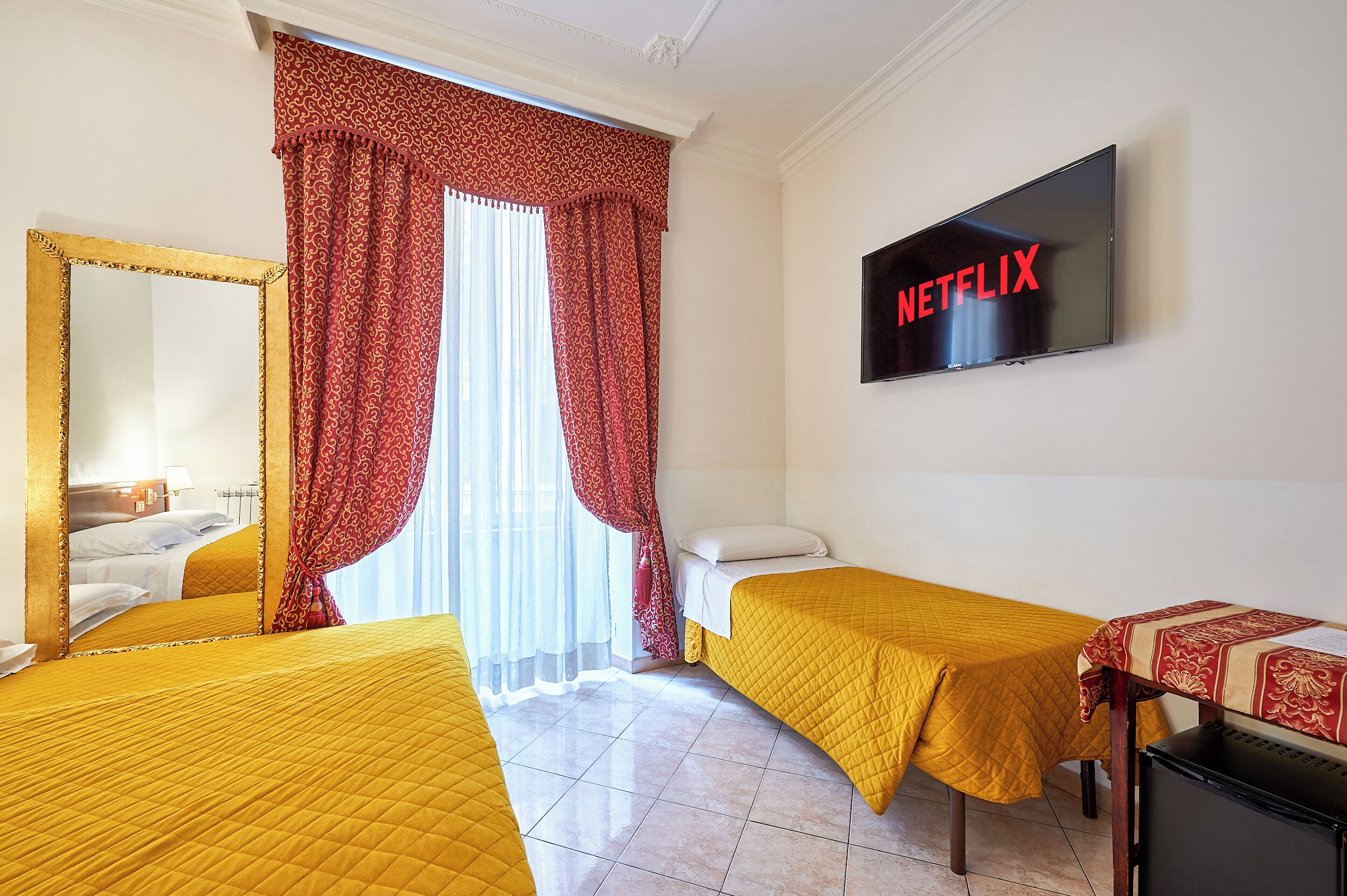 Una camera d'albergo con due letti, una TV a parete e una finestra con tende rosse.