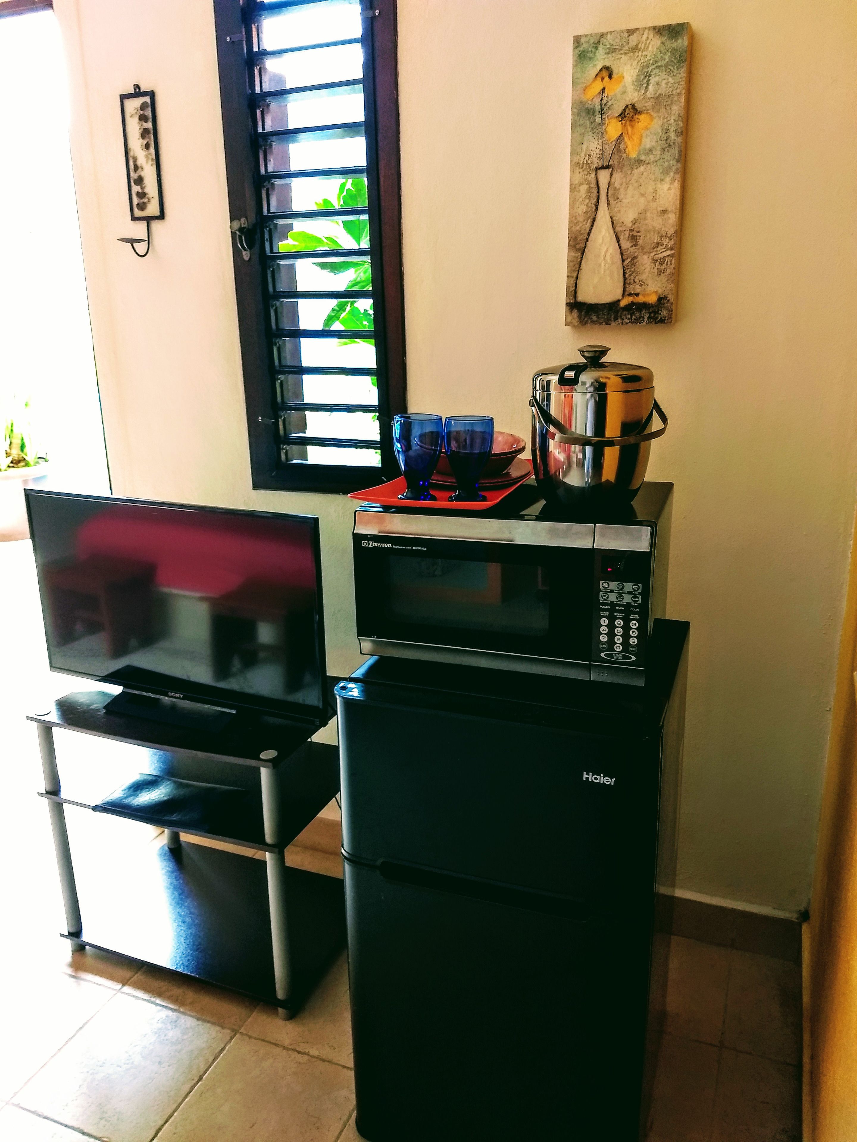 Uma cozinha compacta com micro-ondas, torradeira e um pequeno frigorífico. Uma televisão de tela plana está posicionada em um suporte, e há uma janela com persianas.