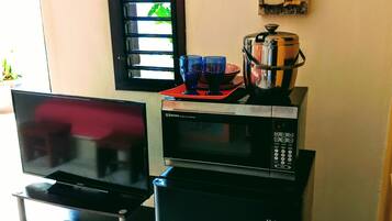 Uma cozinha compacta com micro-ondas, torradeira e um pequeno frigorífico. Uma televisão de tela plana está posicionada em um suporte, e há uma janela com persianas.
