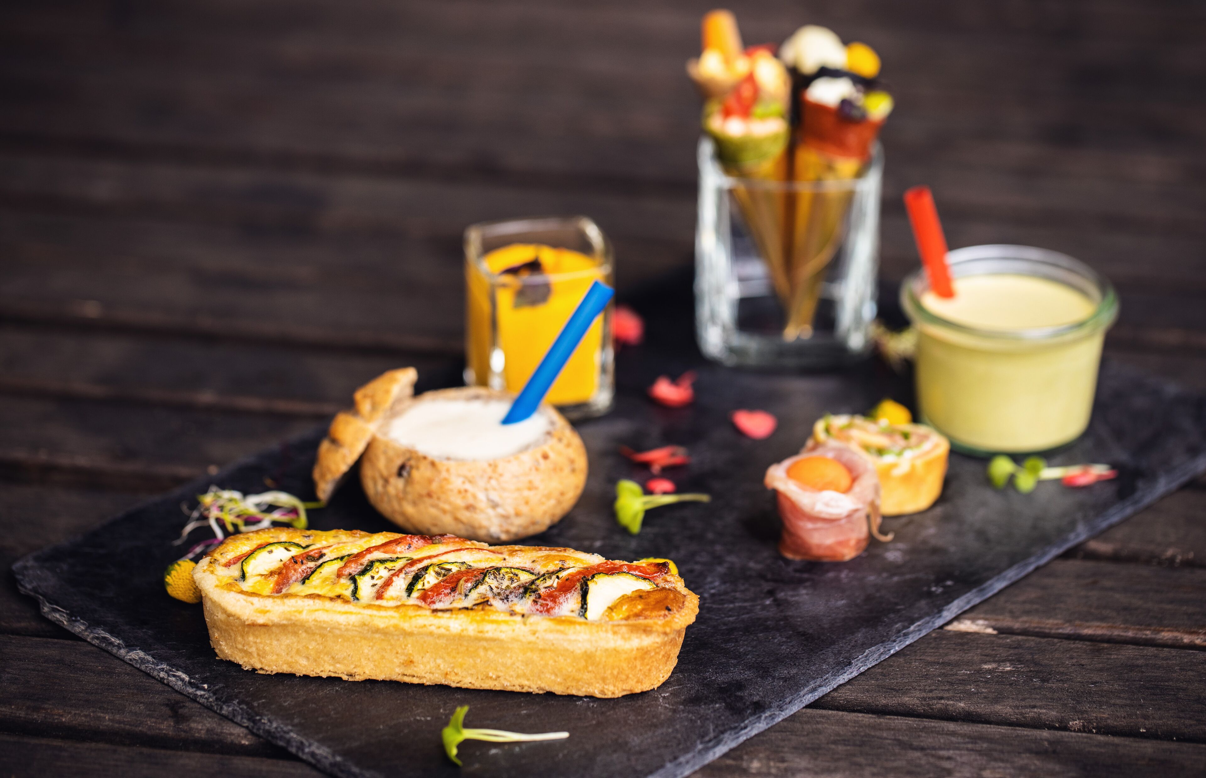A variety of food items on a slate board, including a tart, a bread roll, and a glass of a yellow beverage.