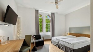 A hotel room with a wooden desk, two beds, a TV, and a window with curtains. - TOP HOTEL ODELYA Basel (Basel)