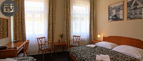 A hotel room with two single beds, a desk with a chair, a small table with a vase of flowers, and two framed pictures on the wall.