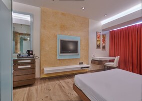 A modern hotel room with a large bed, a desk, a TV mounted on the wall, a mirror, and a red curtain.