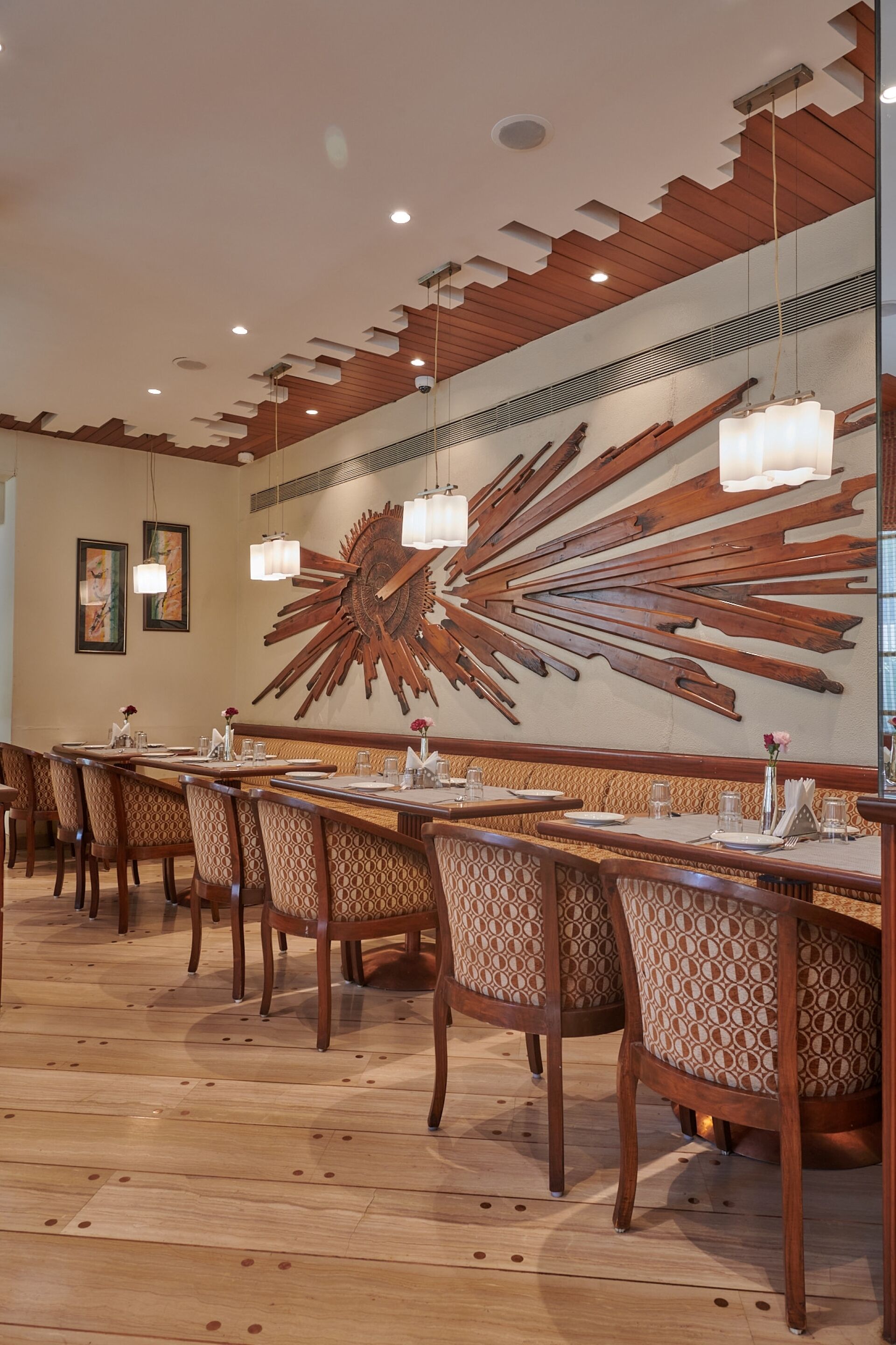A restaurant with wooden tables and chairs, a large decorative wall piece, and pendant lighting.