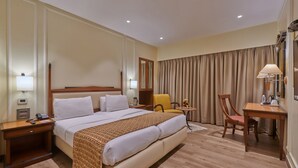 A hotel room with a large bed, bedside tables, a desk with a chair, a lamp, and a window with curtains.