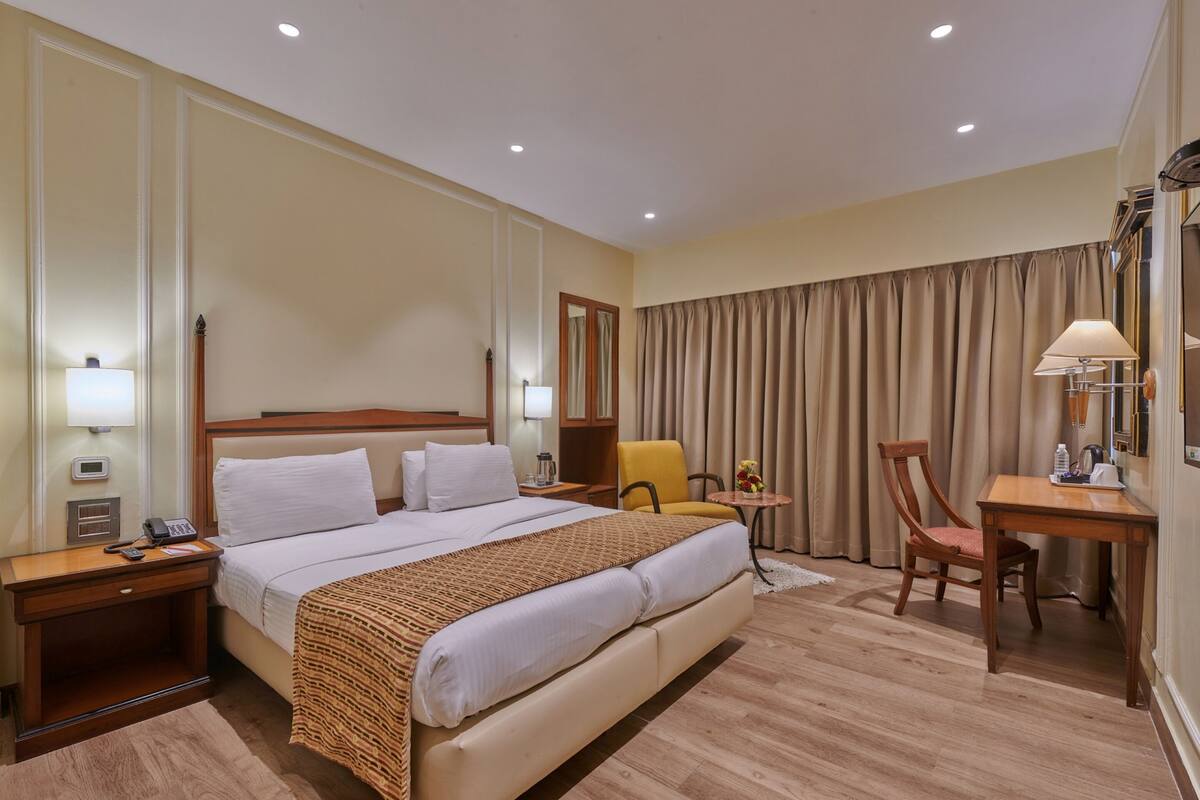Habitación de hotel con una cama grande, mesitas de noche, un escritorio con silla, una lámpara y una ventana con cortinas.
