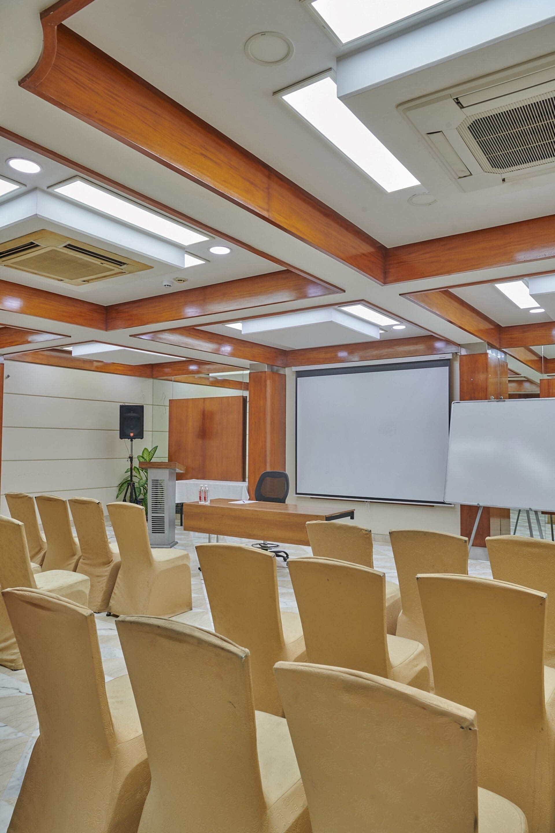 Una sala de conferencias con paneles de madera, una pizarra, una pantalla de proyector y filas de sillas color beige.