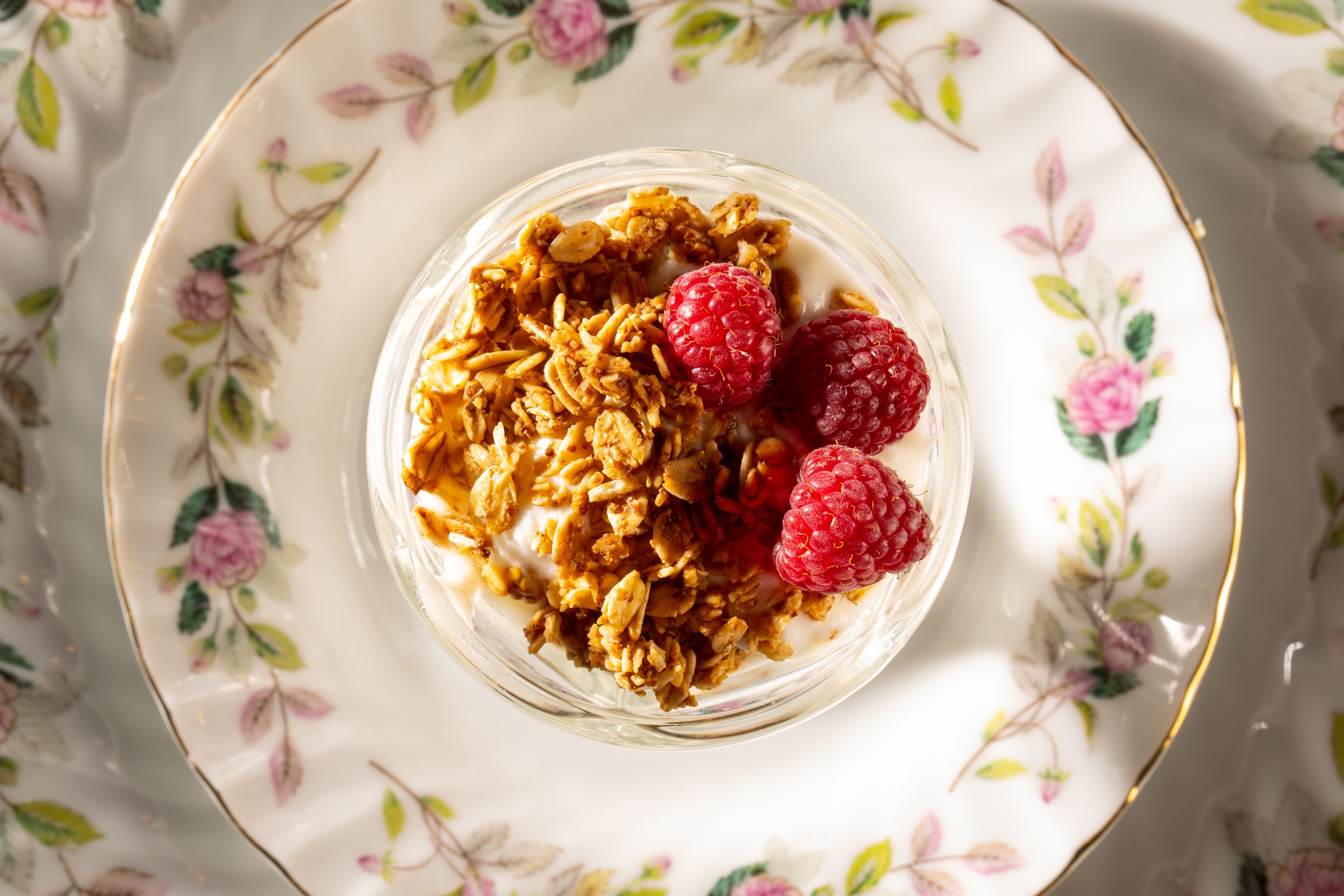 Un bol de granola garni de framboises, présenté sur une assiette à motifs floraux.